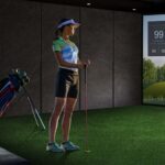 A girl standing in front of a projector in an indoor golf simulator holding a golf club