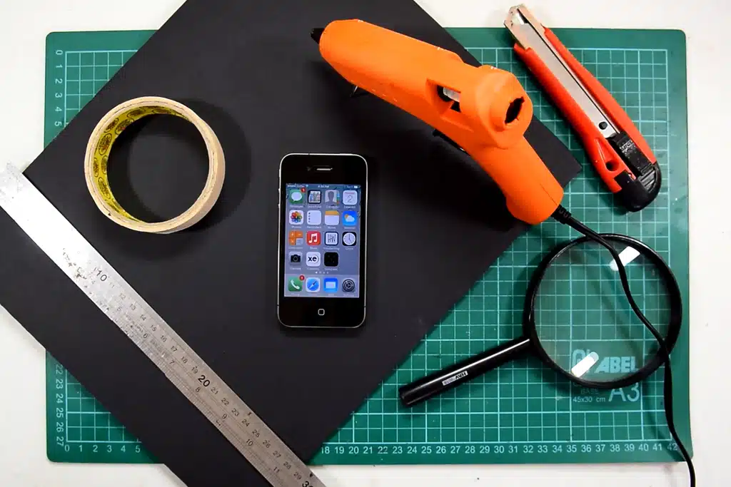 Materials needed for a DIY phone projector including a smartphone magnifying glass glue gun tape ruler and black construction paper