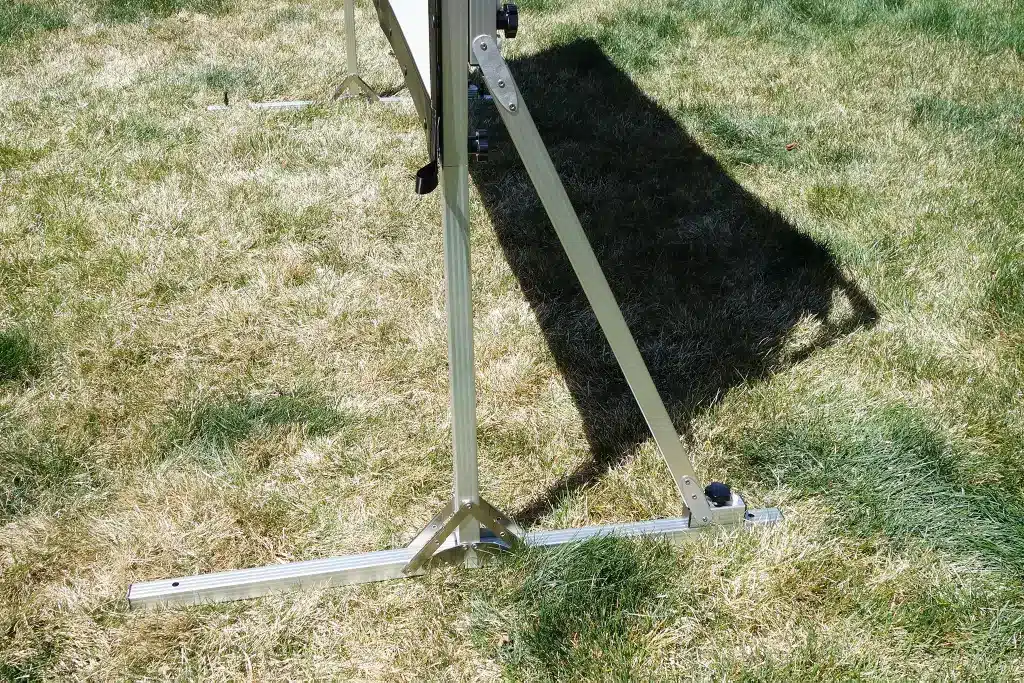 A close up of a projector screen stand set up outdoors on grass showing the sturdy base and support structure