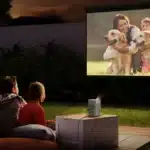 A family watching a movie outdoors with a projector featuring a woman and child with golden retrievers on the screen