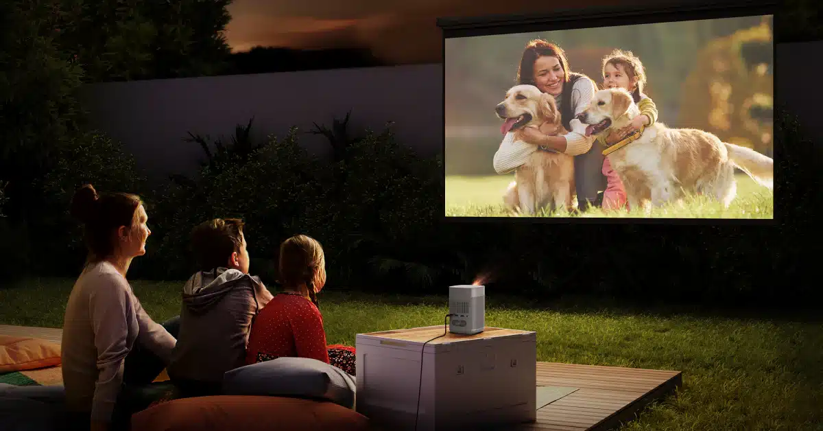 A family watching a movie outdoors with a projector featuring a woman and child with golden retrievers on the screen