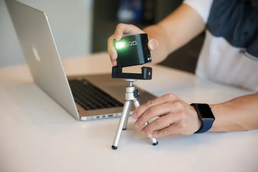 A person setting up a PIQO portable projector on a tripod next to a laptop